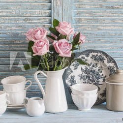 Pink roses in a white enameled pitcher, vintage crockery on blue wooden rustic background. Kitchen still life in vintage style. Flat lay