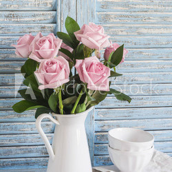 Pink roses in a white enameled pitcher and ceramic white bowls on blue wooden rustic background. Kitchen still life in vintage style