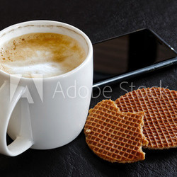 Milky frothy coffee in white mug partially drunk next to partially eaten waffle  biscuits isolated on black.